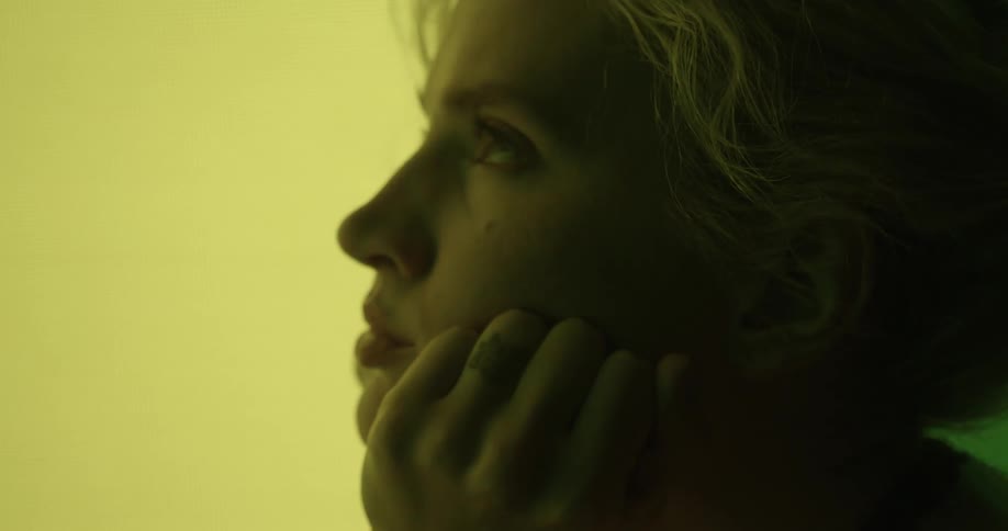 A blonde woman sits with her hands to her chin as the background moves with different lights