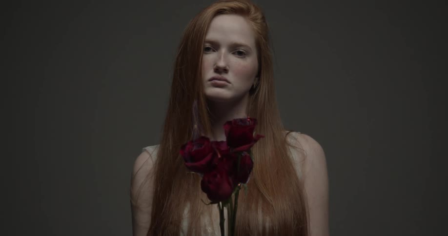 A girl with red hair stands with a stony expression as red roses burn with flames in front of her inside a room