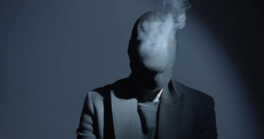 A man poses with a knit beanie pulled down over his head and blows out smoke
