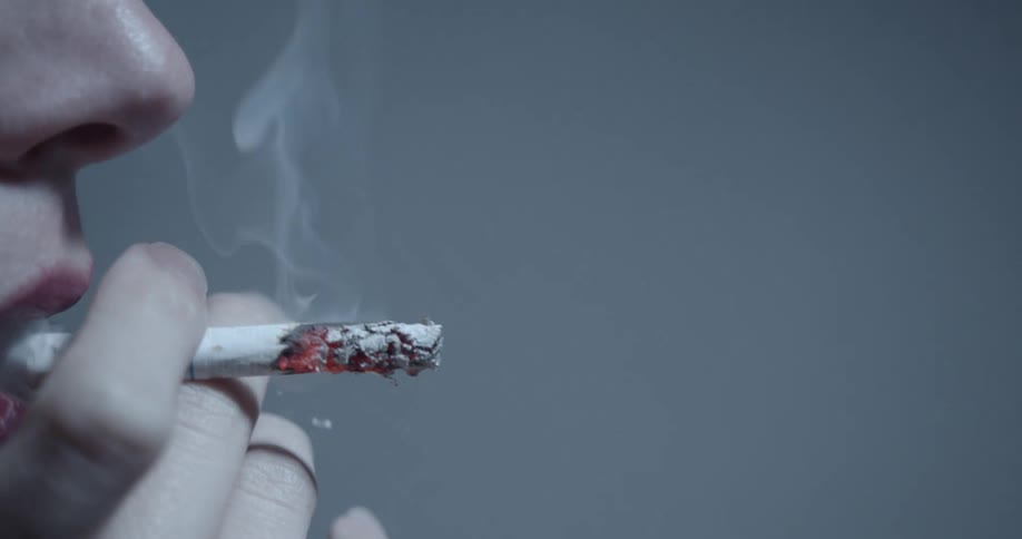 A person takes a drag from a burning cigarette before exhaling the smoke