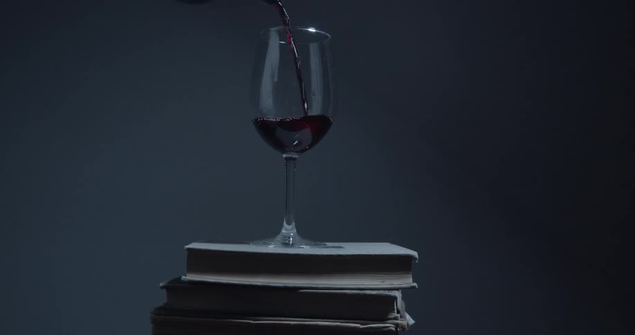 Red wine is being poured into a glass sitting on a stack of dusty and old books
