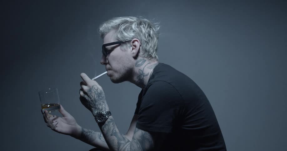 Blonde tattooed man smoking a cigarette and drinking whiskey sitting in a studio