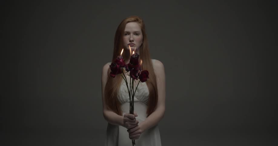 A young woman in a chase holds a bouquet of burning roses while gazing at the camera through the flames