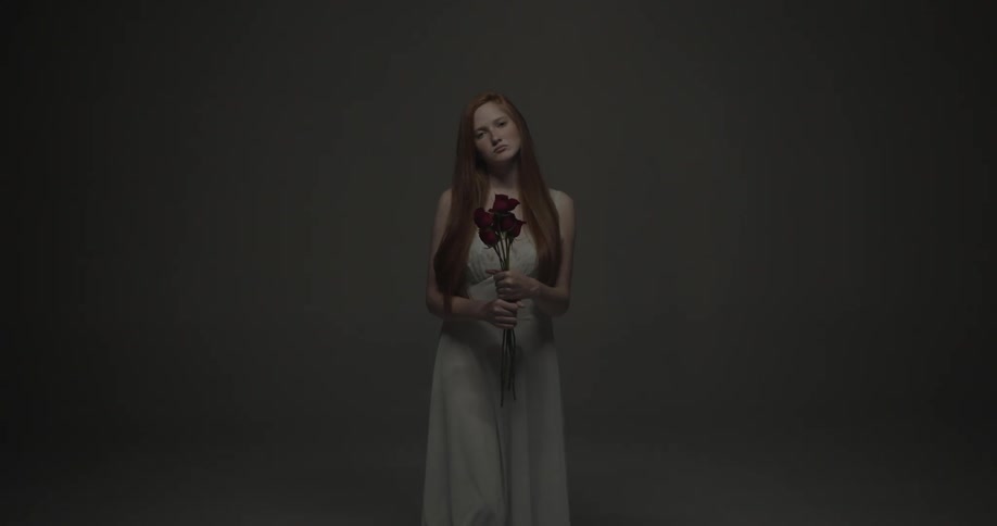 Woman with red hair in white dress stands looking at the camera and holding three roses