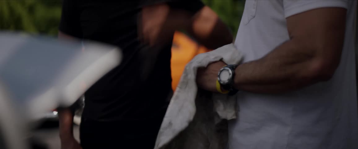 A man wipes off a tool with a dirty towel while he talks to a man in front of a car outside