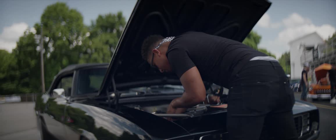 A man works underneath the hood of a car in the parking lot of a car show