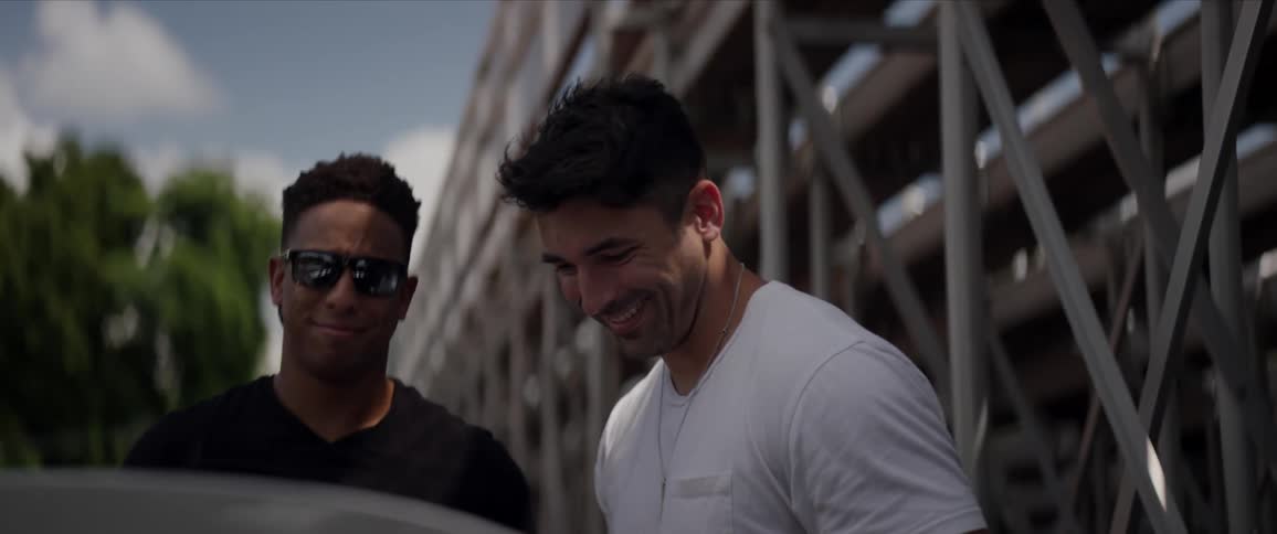 A black male and a white male stand behind bleachers next to a car talking and laughing during the daytime