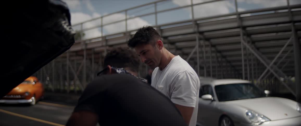 Two man are looking at the engine of a car with the hood open behind bleachers
