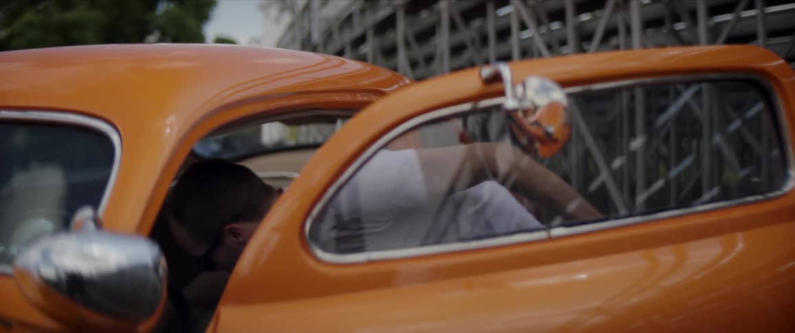 A man wipes down the inside of an orange classic car thats parked behind a stadium with its hood up