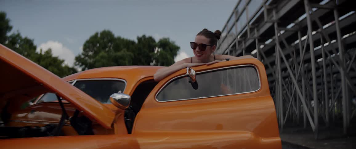 A man walks to the front of a car holding a tool and a woman stands by the drivers side door smiling and talking to him