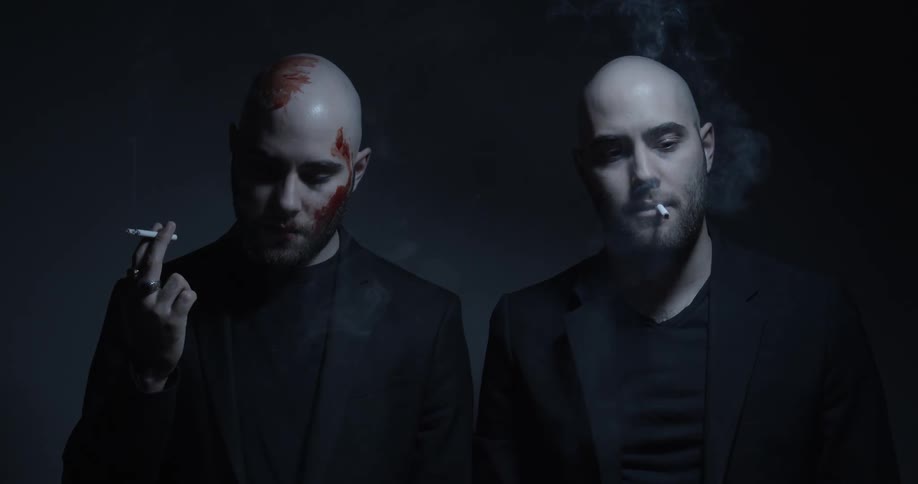 Two bald men covered in blood smoke cigarettes inside a dimly lit room