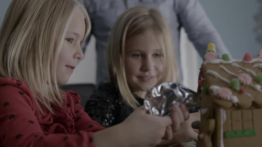 Two young girls decorate a gingerbread house while an adult male watches behind them