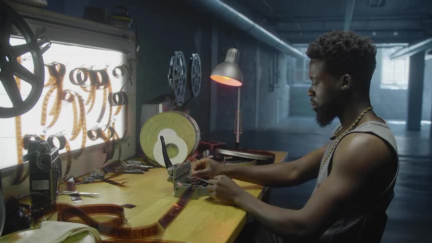 A filmmaker sits at his desk in an open building and uses a tool to trim reels of film