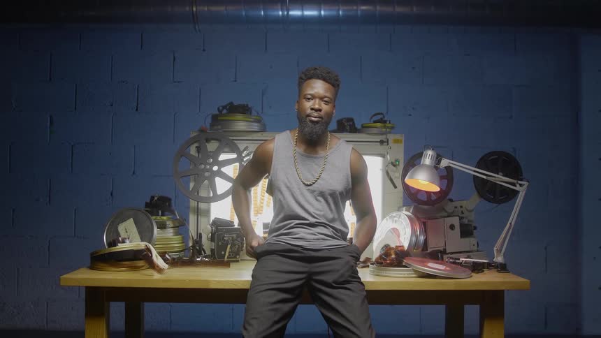 A black man leans against a wooden table against a blue wall