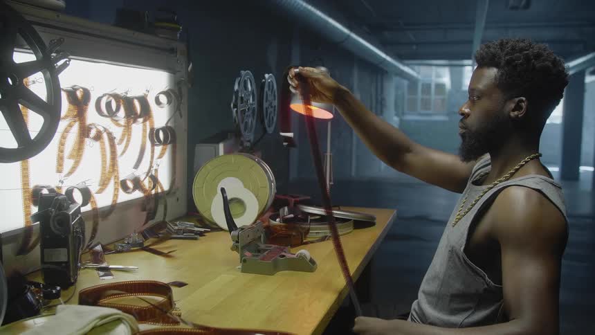 A black man sits at a table holding a piece of film and then cutting it