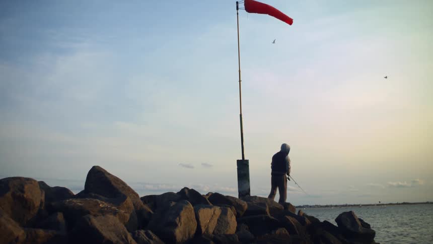 Person wearing hoodie and vest stands fishing on boulders along shore near windsock as birds fly by