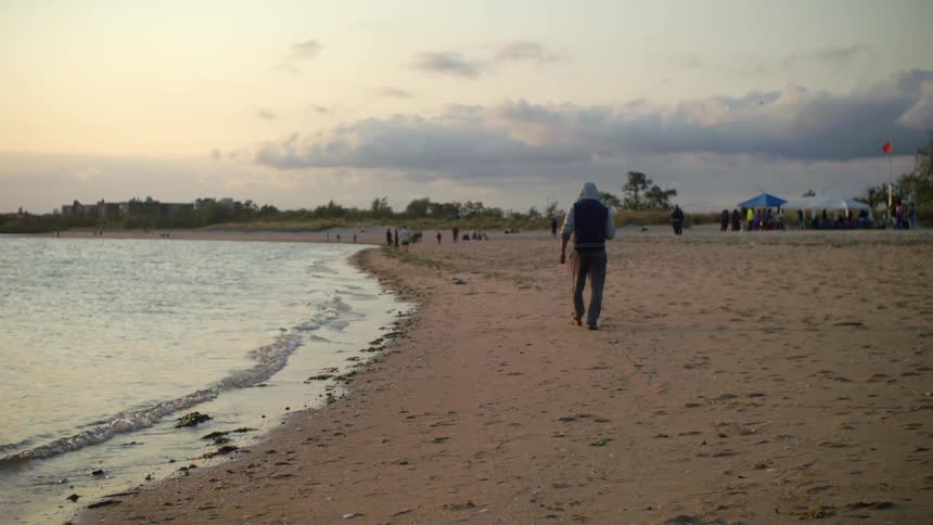 Man wearing a hoodie walking over the sand next to the beach at sunset