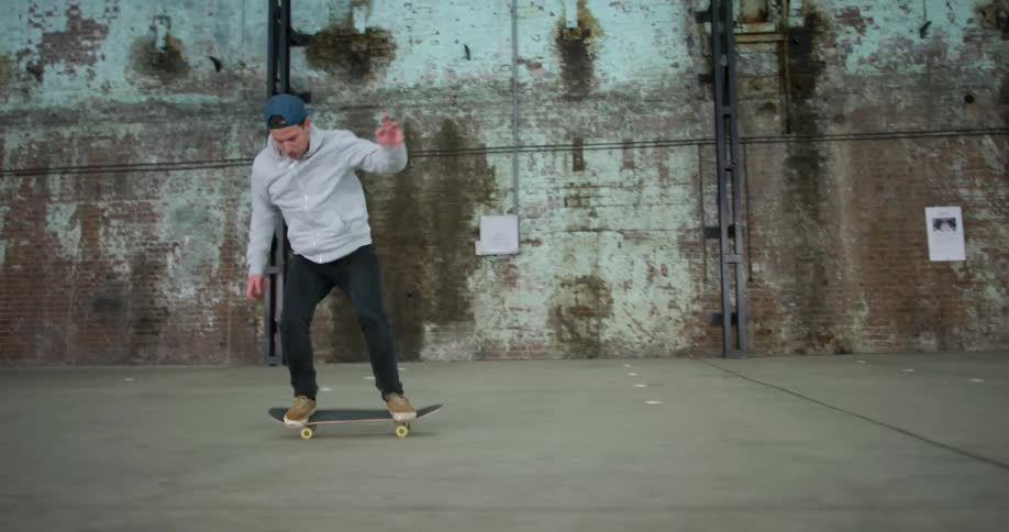 A man with a hat and a hoodie skating on a skateboard with a wall in the background