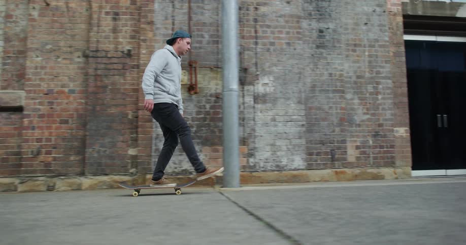 A man skateboards on pavement in front of a brick building with green windows during the day