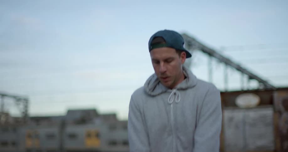 A man wearing a baseball hat and sweatshirt jumps rope in an industrial location with a train running on tracks behind him