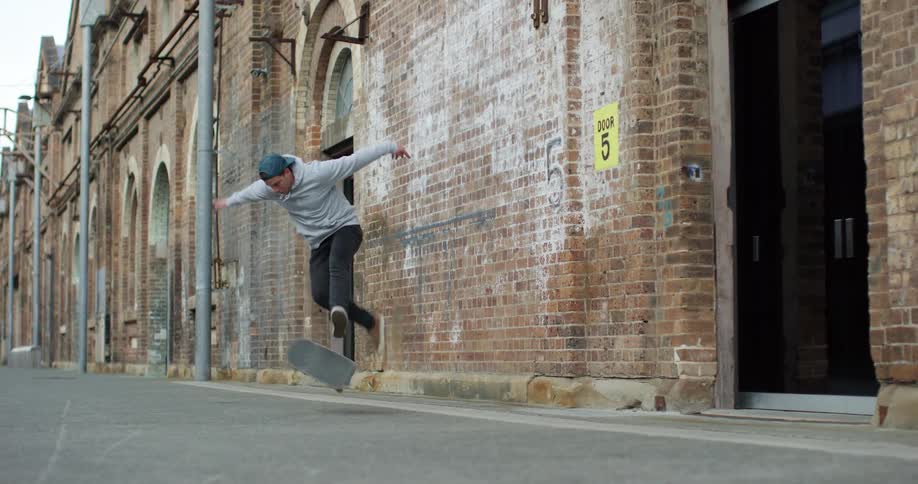 A man wears a backward baseball cap as he rides down a city street doing tricks on a skateboard