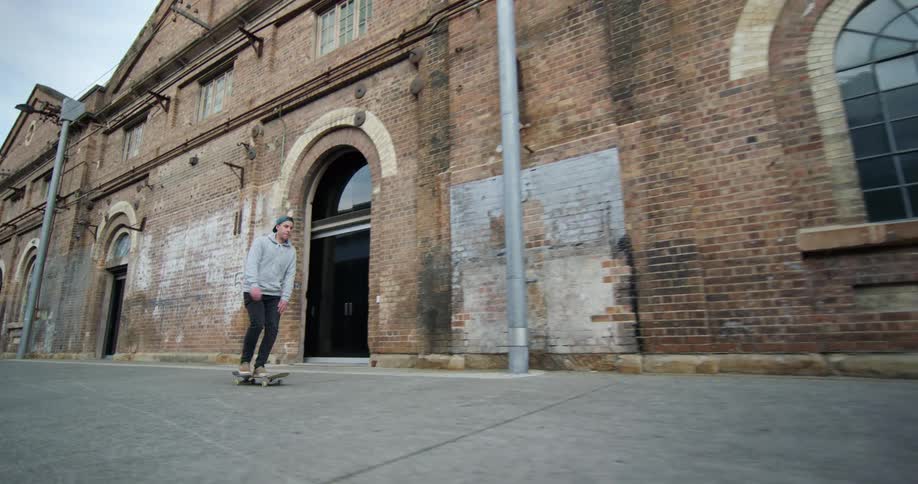 A man rides a skateboard on a roadway beside a long collection of town buildings