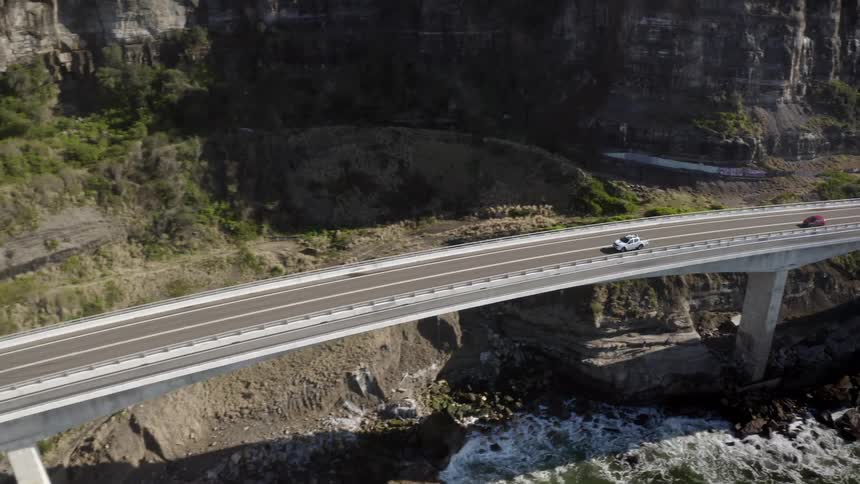 Vehicles drive along a coastal highway as waves crash onto the shore underneath the bridge