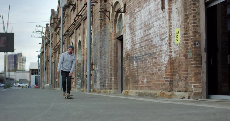 Man with a green cap a grey hoodie and black pants skateboards in the street