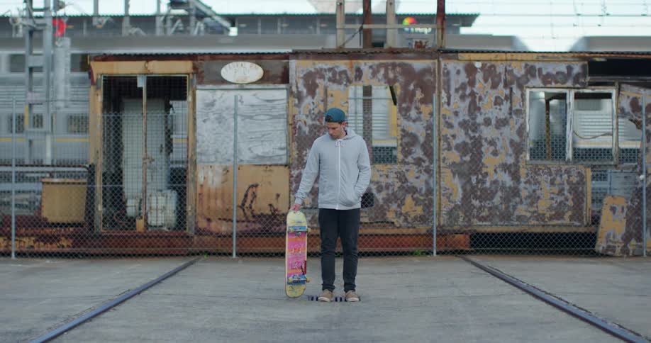 Man with a green cap a grey zip up hoodie and black pants holds a skateboard at the train station