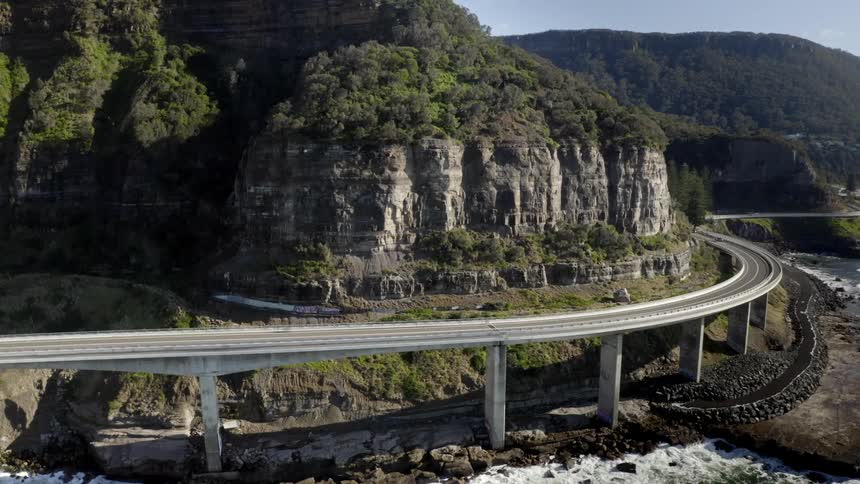 Vehicles travel along a large bridge highway wrapping around a mountainside waterway