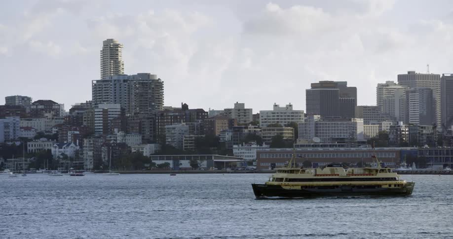 A green and yellow ferry moves along the water with a city at the shoreline on a cloudy day