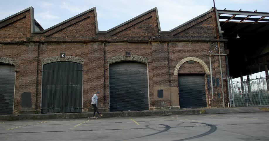 Man leisurely riding a skateboard next to a row of brick warehouses