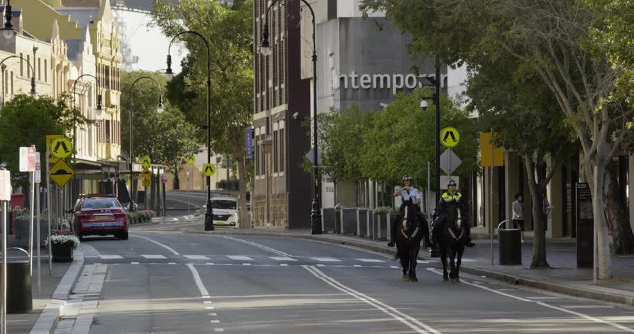 Two police officers riding on two black horses down a street in a small town while pedestrians walk by on the right