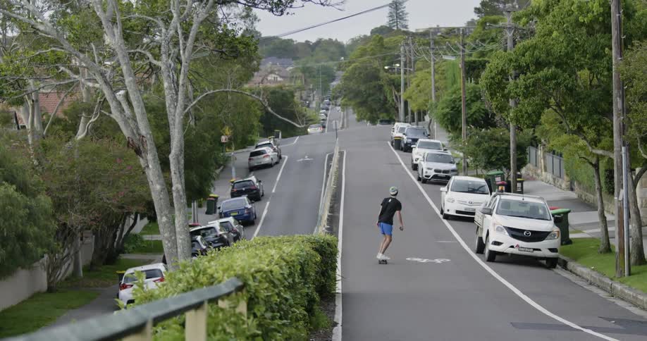 A man with a hat skateboarding on a road with parked vehicles with trees the sky and clouds in the background