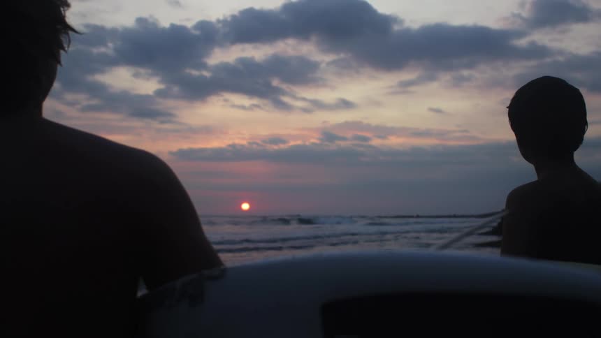 Man and boy holding surfboards on the beach are silhouetted against the sunset