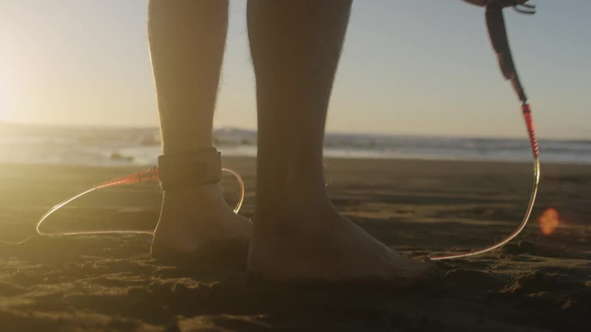 Surfer standing on a beach at sunset lifting his surfboard off of the ground in front of the ocean