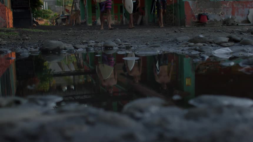 A close up of a rocky ground with a puddle and three people walking and holding surfboards