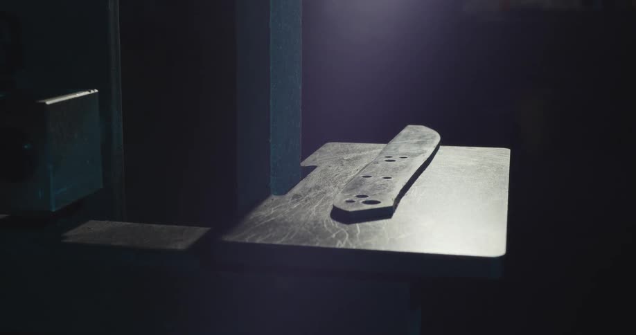 Flare of light shining down on the blade of an unfinished knife on a machine