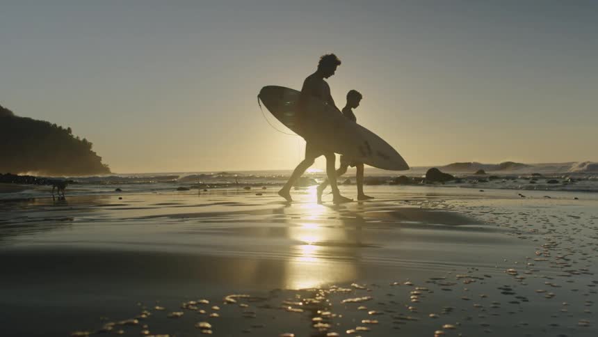 A pair of surfers carry surfboards as they walk across a beach under a low sun