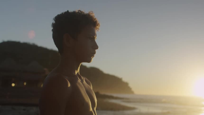 Teenage boy watches the sea on the beach at sunset