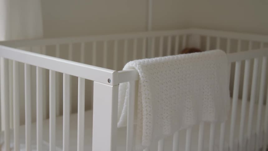 A white crib sits in a room next to a window with a white blanket draped over the railing