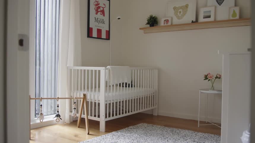 A babys nursery with a crib and a rug there are pictures on a shelf with a plant and another picture on the wall there are flowers on a white table and a toy in the floor beside the window with blue curtains