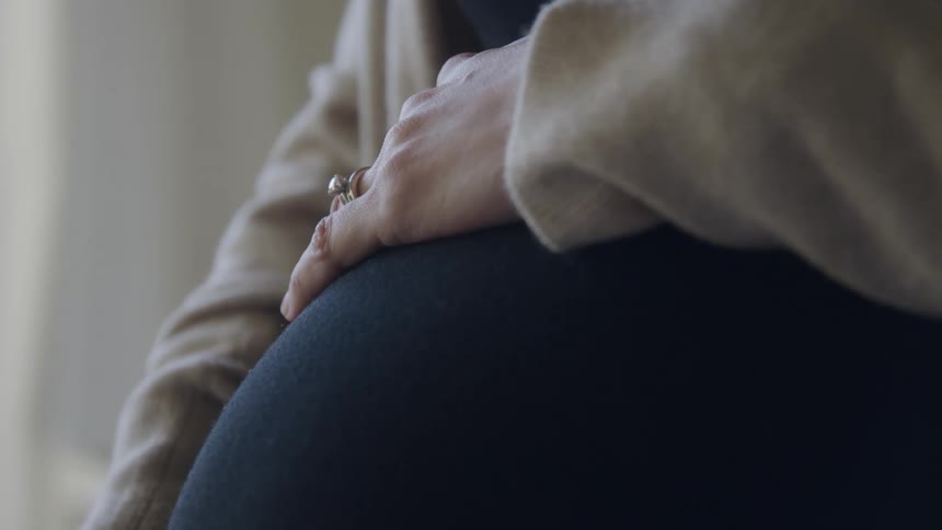 Pregnant woman with a black sweatshirt and rings touches her belly in a room