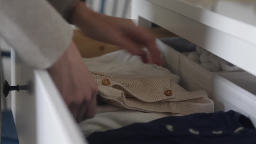 Pregnant woman storing folded baby clothes in a dresser drawer on a room