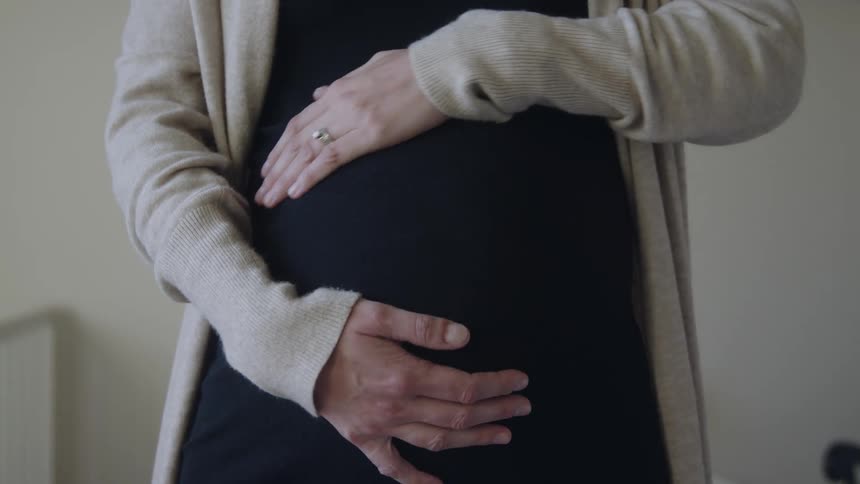 Pregnant woman with a black sweatshirt and rings touches her belly in a room