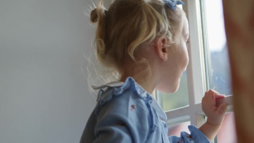 Blonde little girl with a blue dress looks out the window in a room