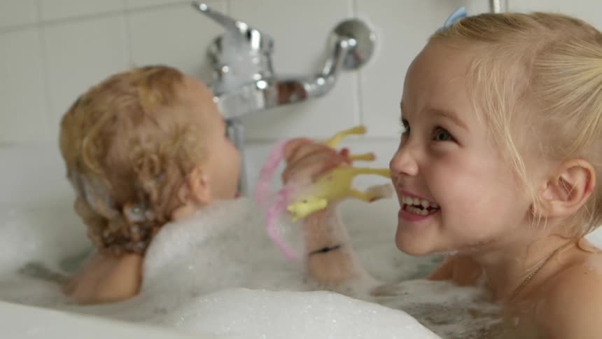 Blonde girls sit in bathtub filled with water and enjoy themselves in the bathroom