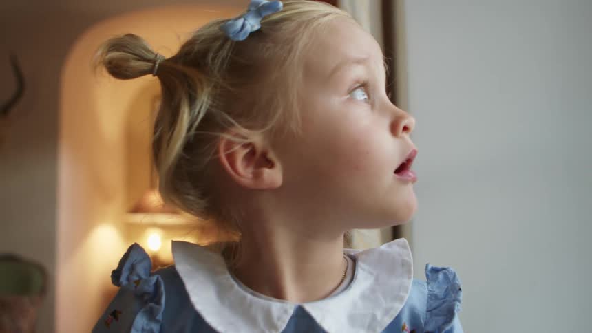 A blonde toddler sits in her house and looks back and forth with wide eyes
