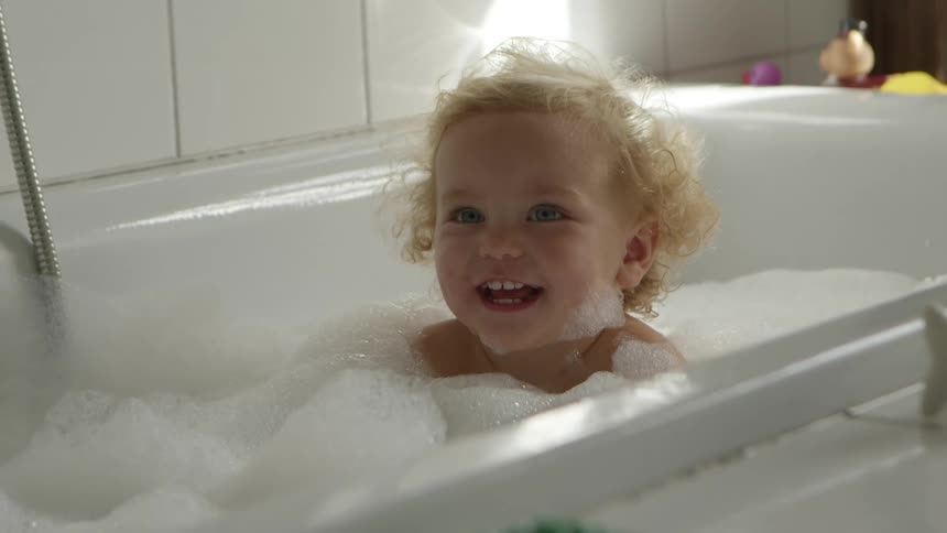 Blonde toddler girl sits in bathtub filled with water in the bathroom
