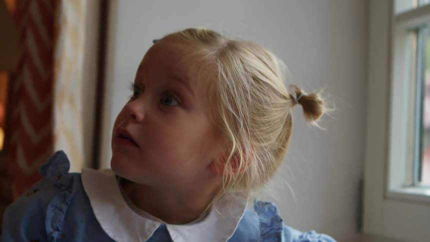 Young blonde girl in a blue dress looks excitedly through a window at home